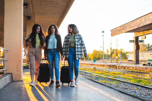 istock Three beautiful young female friends walking at a train station with two suitcases during sunset while waiting to travel by train 1366147774