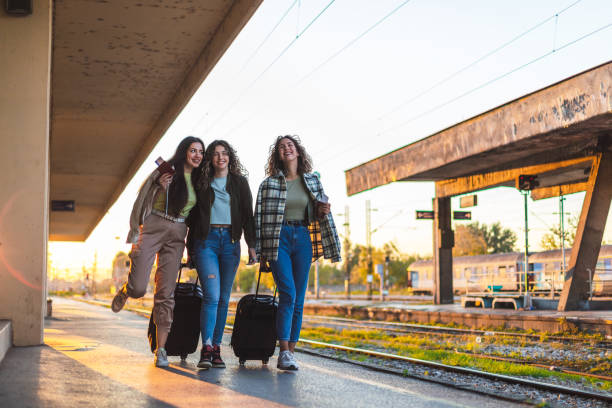 istock Three beautiful young female friends walking at a train station with two suitcases during sunset while waiting to travel by train 1366147781