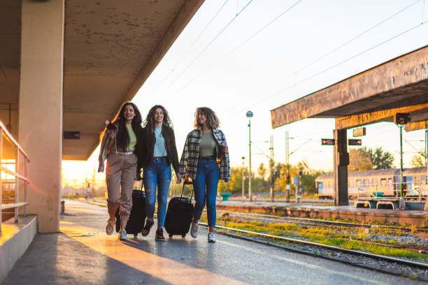 istock Three beautiful young female friends walking at a train station with two suitcases during sunset while waiting to travel by train 1366147782