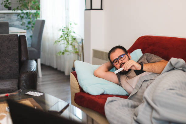 istock Young man with glasses sick with COVID-19 checking his fever temperature while laying in bed at home 1366147923