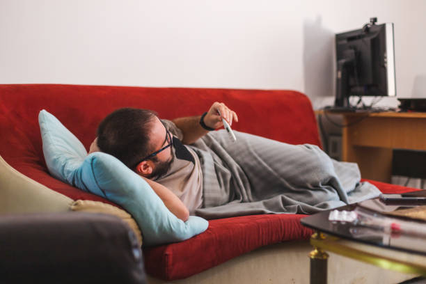 istock Young man with glasses sick with COVID-19 checking his fever temperature while laying in bed at home 1366147929