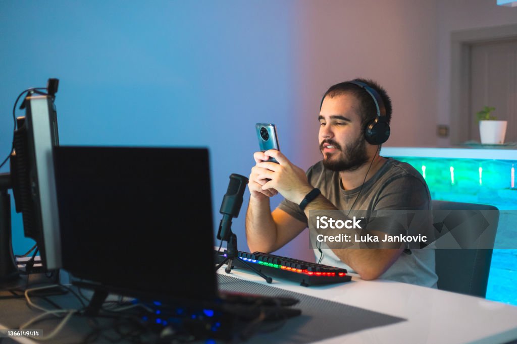 Happy young male gamer using his mobile smart phone while streaming with a keyboard and a microphone in front of him Young male gamer streaming while working from home. Gamer Stock Photo