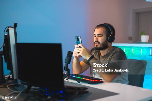 Happy Young Male Gamer Using His Mobile Smart Phone While Streaming With A Keyboard And A Microphone In Front Of Him Stock Photo - Download Image Now