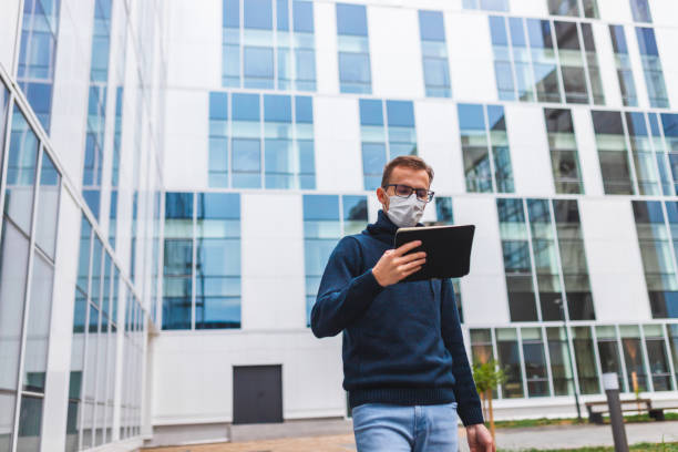 istock Young man wearing glasses and a protective face mask pulling his suitcase outside an airport while looking at his tablet 1366150065