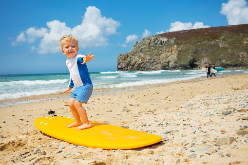 Little blond toddler boy already practicing and posing balancing standing on the surfboard on the beach