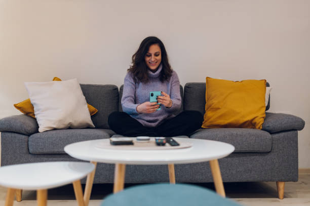 istock Hispanic woman using smartphone while sitting on the couch at home 1366158078