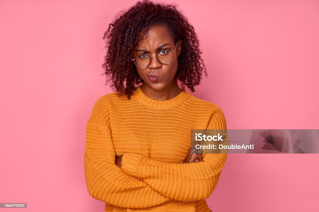 Sad young African American woman displeased looking at camera with crossed arms over pink background Sad angry young african american woman displeased looking at the camera with her arms crossed over a pink background in the studio. Concept of failure. Advertising space Displeased Stock Photo