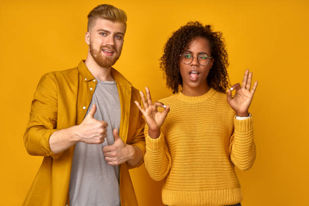 excited beautiful mixed race couple shows thumb up gesture agree to do something and collaborate, in yellow clothes against orange wall, did great job, show approval, like idea. everything will be ok - thumbs up business people isolated imagens e fotografias de stock