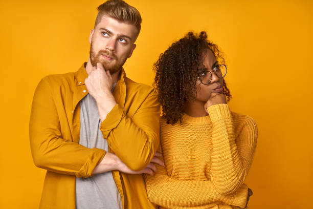 headshot of puzzled ginger husband and his african american wife hold chin and purse lips, looks with hesitation at camera, think what place for vacation to choose. young family couple stand indoor - vacations two generation family caucasian friendship imagens e fotografias de stock