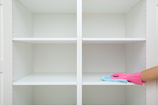 Young adult woman hand in pink rubber protective glove using blue dry rag and wiping white shelves inside wooden wardrobe in room. Closeup. Front view. Regular cleanup at home.