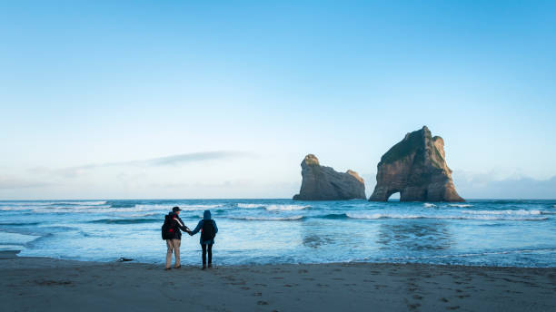 casal em pé na praia wharariki e assistindo archway island em sunrise, ilha sul da nova zelândia. - sunrise beach couple hiking - fotografias e filmes do acervo