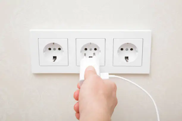 Photo of Young adult woman hand holding and plugging white electrical plug for smartphone charging in wall outlet socket at home. Closeup. Point of view shot.