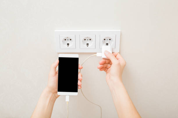 giovane donna adulta che tiene la mano e collega la spina elettrica bianca per la ricarica dello smartphone nella presa a muro a casa. primo piano. punto di vista ripreso. posto vuoto per il testo sullo schermo nero. - mobile phone charging power plug adapter foto e immagini stock