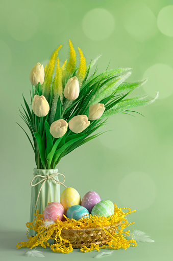 Multi colored hand-painted Easter eggs in a basket with the bouquet of tulips in a vase. Defocused lights background with beautiful bokeh. Space for copy.