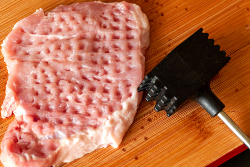 Raw meat slice and iron meat tenderizer on wooden cutting board.