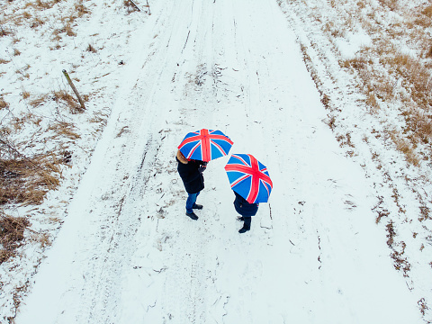 Aerial image taken by drone depicting a couple walking in a freezing rural landscape covered with snow in the middle of winter. They are holding identical umbrellas with a British union jack flag design. Lots of room for copy space.