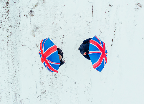 Aerial image taken by drone depicting a couple walking in a freezing rural landscape covered with snow in the middle of winter. They are holding identical umbrellas with a British union jack flag design. Lots of room for copy space.