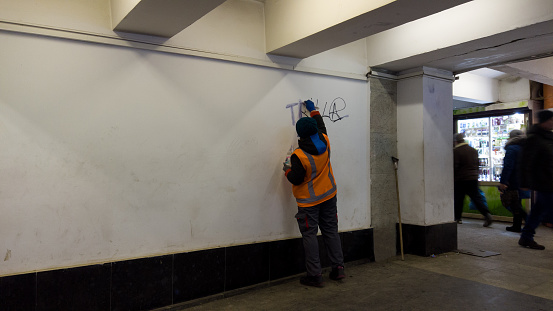 Removing graffiti from vandals in underground pass. employee of the city, wearing a orange uniform, cleans wall from graffiti and writing, with a liquid solvent. Kharkiv, Ukraine - December 27, 2022