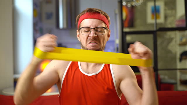 Portrait of funny man in red retro sportswear doing exercises with an elastic resistance band at home.