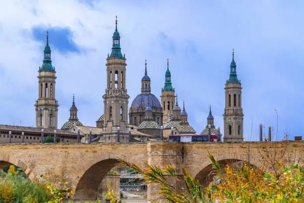 Medieval stone bridge Puente de Piedra with arches on the background of the belltowers of the Basilica of Our Lady of the Pillar in Saragossa. Ancient architecture of a Spanish town