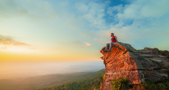 man on the cliff,Young man tourist with backpack relaxing on top rock and enjoying sunset,Young Man sit on the peak of great rock.