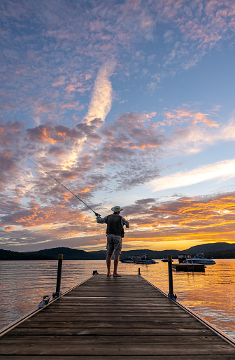 Active Senior Man Fly-Fishing at Sunset at the lake in summer. He is standing on a pier at Lac St-Joseph, Quebec, Canada. The sky is multi colored with orange, red, yellow and purple colors. It is a romantic sky.