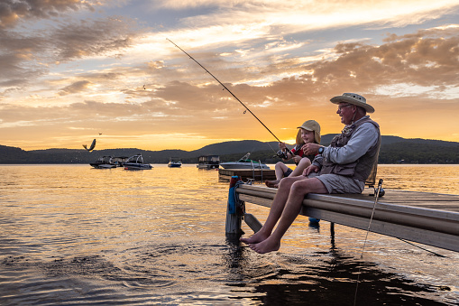 A grandfather is teaching his grandson to fish during sunset in summer. They are both sitting on the dock, pier, and laughing. It is a beautiful summer day. The little boy just caught a fish. Across the lake, there is a mountain. Lac Saint-Joseph, Fossambault-sur-le-Lac, Quebec, Canada.