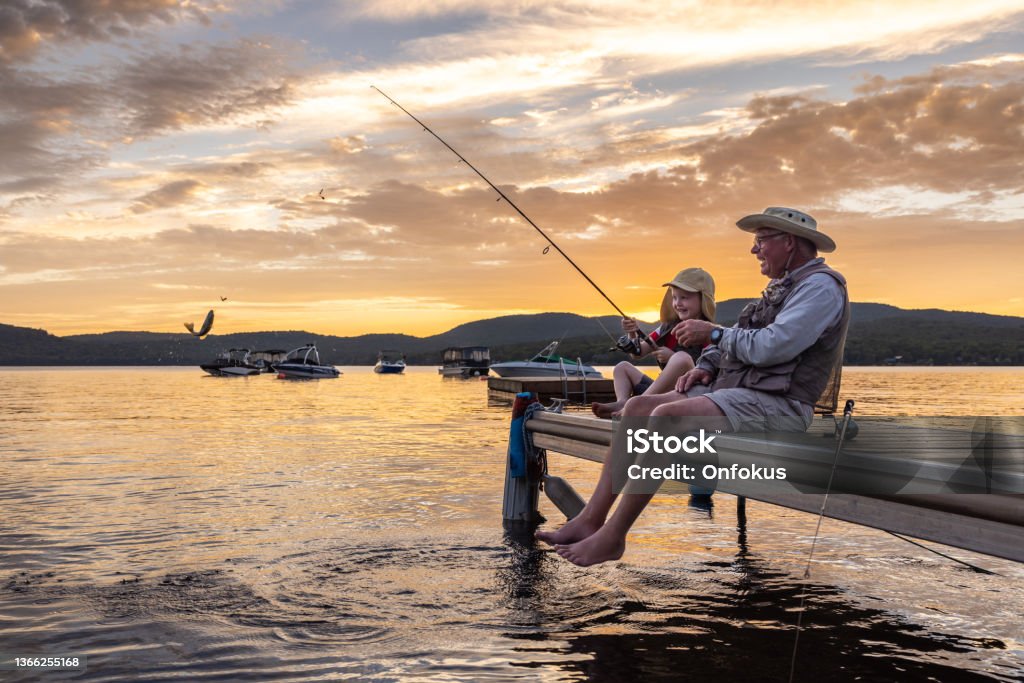 Grandfather and Grandson Fishing At Sunset in Summer, Quebec, Canada A grandfather is teaching his grandson to fish during sunset in summer. They are both sitting on the dock, pier, and laughing. It is a beautiful summer day. The little boy just caught a fish. Across the lake, there is a mountain. Lac Saint-Joseph, Fossambault-sur-le-Lac, Quebec, Canada. Fishing Stock Photo