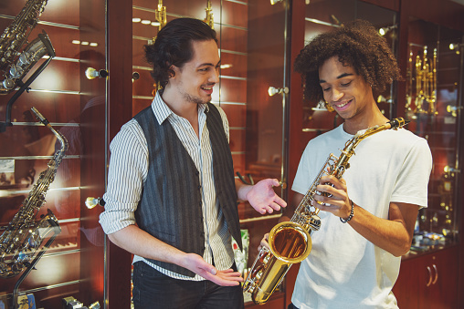 Two handsome stylish men holding a saxophone, talking and smiling while standing in a musical shop