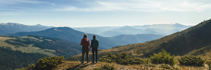 Couple of hikers with backpacks enjoying valley landscape view from top of a mountain. Young adult tourists, man and woman standing on the pass and holding by hands. Panoramic view of mountain hills, Carpathian mountains landscape.