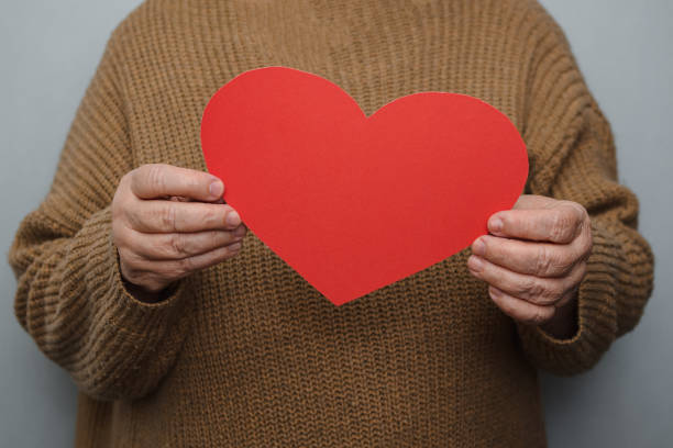 Against the background of a gray wall, selective focus on the hands of an elderly woman, dressed in a warm knitted jacket, holding a large homemade heart cut out of red cardboard at chest level, showing that she loves and is ready to support in difficult times, space for text.