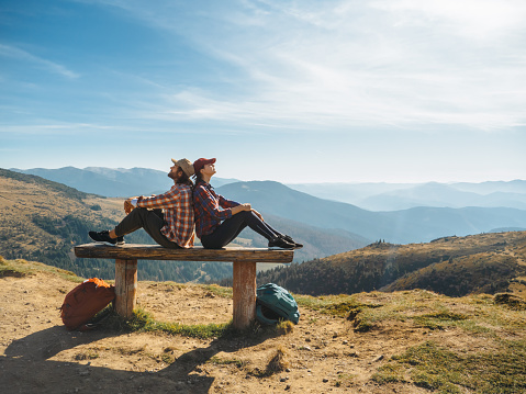 Couple of hikers with backpacks enjoying valley landscape view from top of a mountain.Happy young adult tourists, man and woman sitting back to back on the bench and looking away. Panoramic view of mountain hills, Carpathian mountains landscape.