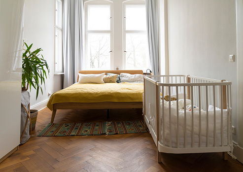 Bedroom of a family with double bed and baby crib. kid's cradle in bedroom interior with large windows and hardwood flooring.