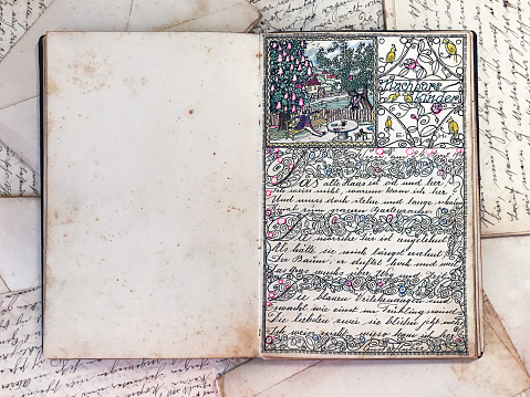 Overhead view of a vintage journal open-wide with a full-page decoration,  on top of old scattered letters from the beginning of the 20th century.