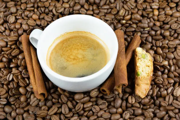 A coffee in white cup surrounded by coffee beans, cinnamon sticks and a bisquit