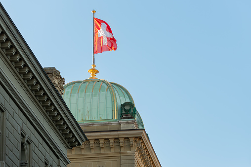 the flag waving in the wind above one of the domes of the Bundeshaus building. 02/21/2021 - Bundesplatz, 3011 Bern, Canton Bern, Switzerland