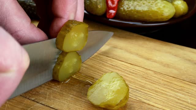 Marinated pickled cucumber are cutting on wooden kitchen board with sharp knife.