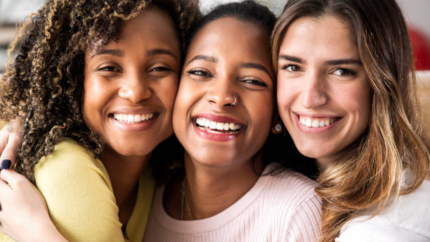 três lindas amigas sorridentes abraçadas - grupo multirracial de mulheres olhando para a câmera - mulheres e conceito de estilo de vida feliz - young women friendship smiling individuality - fotografias e filmes do acervo