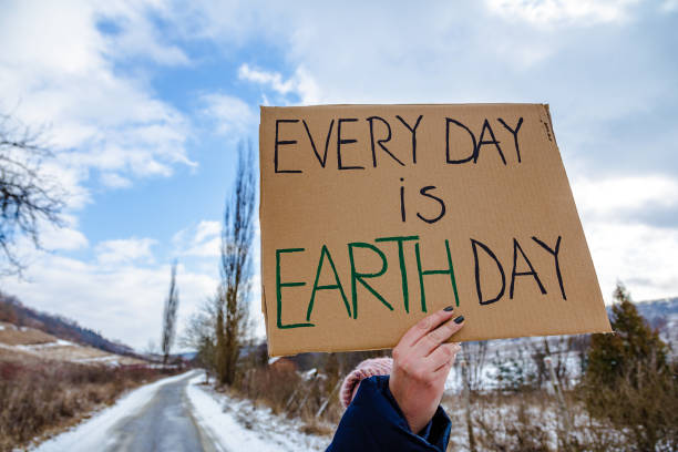 A mid-adult female activist holding a sign protesting against climate change and global warming. She is standing outdoors on a country road in the snow and is holding signs with various messages, such as 'act now' and 'no planet b'. Focus on the sign, with the road and countryside defocused beyond. Room for copy space.