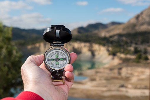 Hand of a hiker holding a compass to orient himself in a westerly direction. In the background is a river, blue sky and mountains out of focus.
