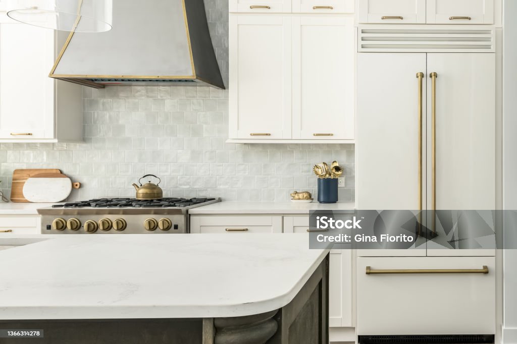 Contemporary White Kitchen with White Matte Appliances and Gold Fixtures Elegant Kitchen Design with white matte refrigerator, white cabinets, and two toned kitchen island. Gray tile backsplash and metal range hood. Kitchen Stock Photo