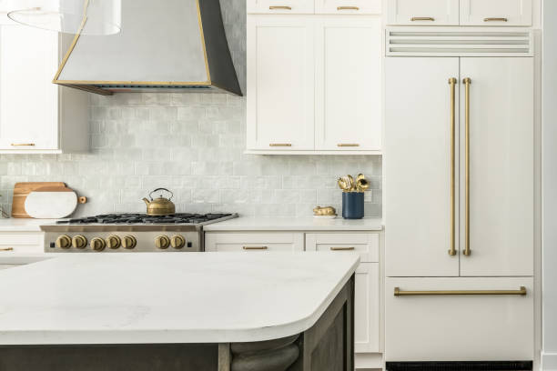 Contemporary White Kitchen with White Matte Appliances and Gold Fixtures Elegant Kitchen Design with white matte refrigerator, white cabinets, and two toned kitchen island. Gray tile backsplash and metal range hood. kitchen stock pictures, royalty-free photos & images