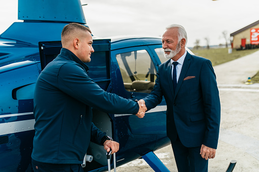 Handsome wealthy senior businessman standing with pilot on airport and using his own air transport chopper or helicopter to travel on business meeting.