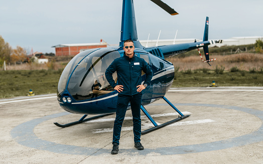 Handsome young adult pilot standing and posing in front of a small private business chopper or helicopter.