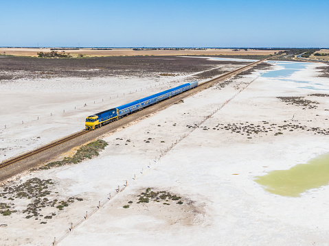 Aerial view Overland Passenger Train crossing dry salt lake on a raised stone causeway with remnant water pools. Train is heading towards the camera en route to Melbourne with yellow farmlands and low hills in the distance. ID & logos edited