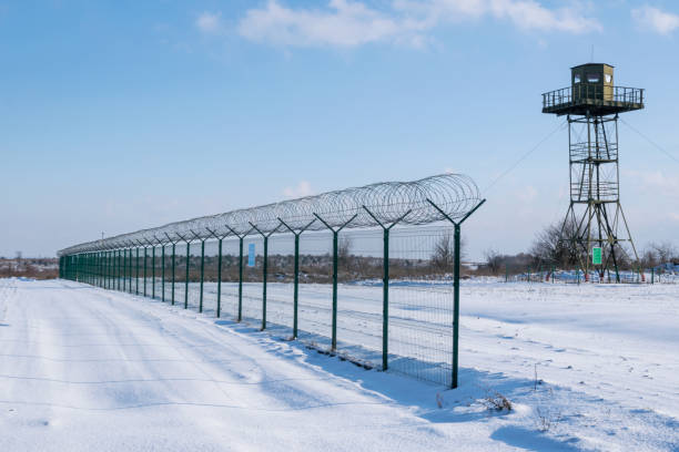 clôture frontalière avec des barbelés devant la tour de surveillance dans un champ enneigé contre un ciel bleu. concept de protection des frontières et de lutte contre la migration illégale - winter wire barbed wire protection photos et images de collection