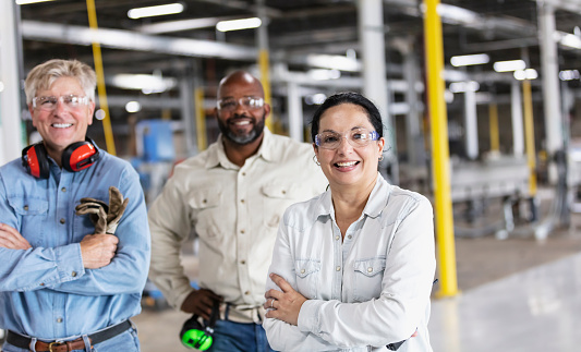 A multi-ethnic group of three workers, a woman and two men, in a plastics factory, standing on the factory floor, looking at the camera. The focus is on the woman, who is Hispanic, in her 40s, smiling with her arms crossed.