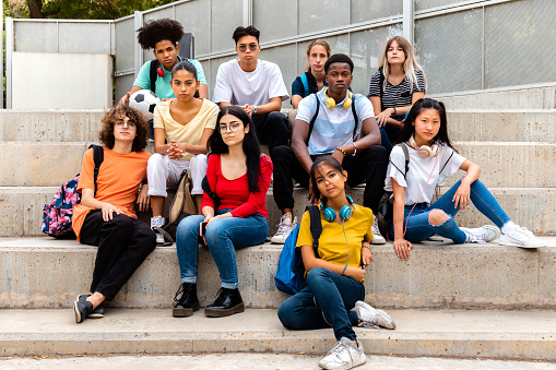 Multiracial teenage students sitting on steps in high school outside looking at camera with serious expression. Education concept.