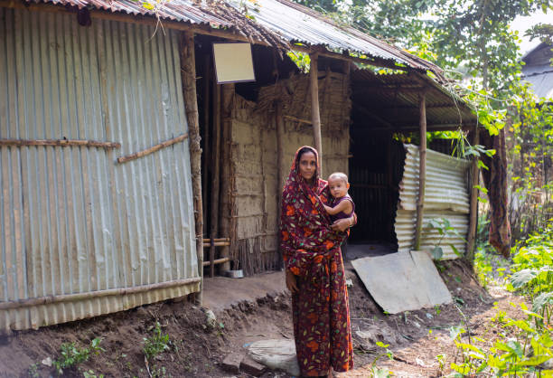 Malnutrition women and child standing in front of the broken bamboo, fence and tin sheet house, and homestead garden planted by her. Gowainghat, Bangladesh - November 06, 2019: Malnutrition woman and child standing in front of the broken bamboo, fence, tin sheet house, and homestead garden planted by her. family mother poverty sadness stock pictures, royalty-free photos & images