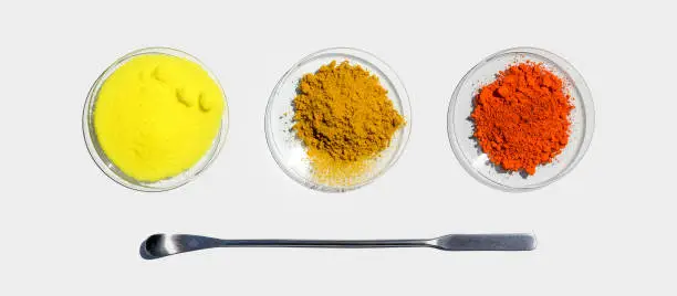 Photo of Lead (II,IV) oxide, Potassium Chromate and Organic Curcuma Powder in Chemical Watch Glass placed next to the stainless spatula. Closeup chemical ingredient on white laboratory table. Top View